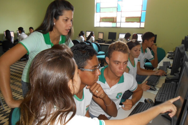 Quatro estudantes estão apontando para um computador em uma sala com outros estudantes