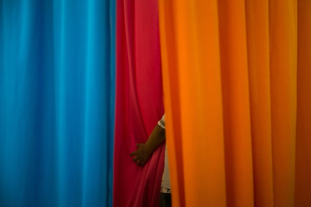 Criança se esconde atrás de tecidos coloridos. - Foto: Nicolas Neves dos Santos