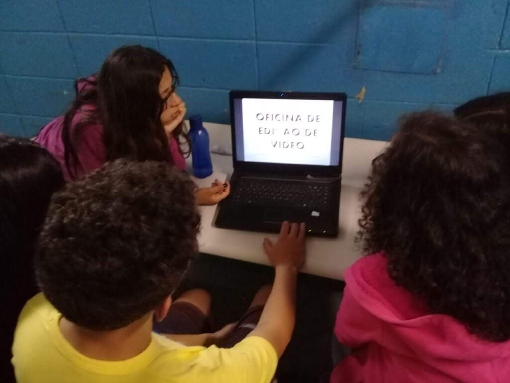 Foto de três jovens sentados à mesa em frente a um notebook. Na tela do computador, está escrito "Oficina de Edição de Vídeo". Os jovens estão em um ambiente fechado com uma parede azul à sua frente.