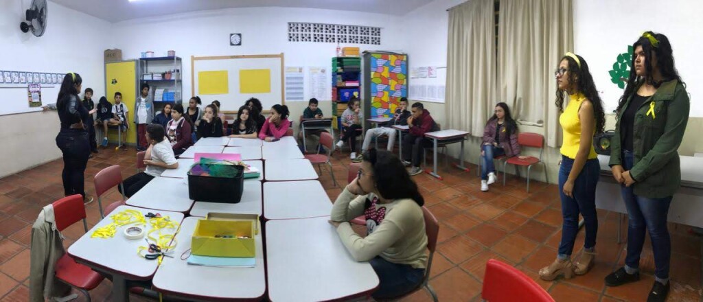 Foto de uma sala de aula com estudantes sentados ao fundo. A professora está em pé à esquerda. No centro da imagem, há carteiras enfileiradas. À direita da foto e ao fundo, há um armário e mais duas estudantes sentadas e duas em pé.