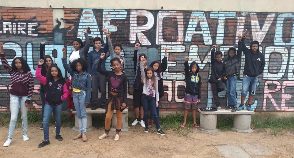 Foto onde há 15 alunos posando em uma área externa na frente de um muro onde se lê AFROATIVOS. Os alunos estão em pé, um ao lado do outro e de punhos cerrados.