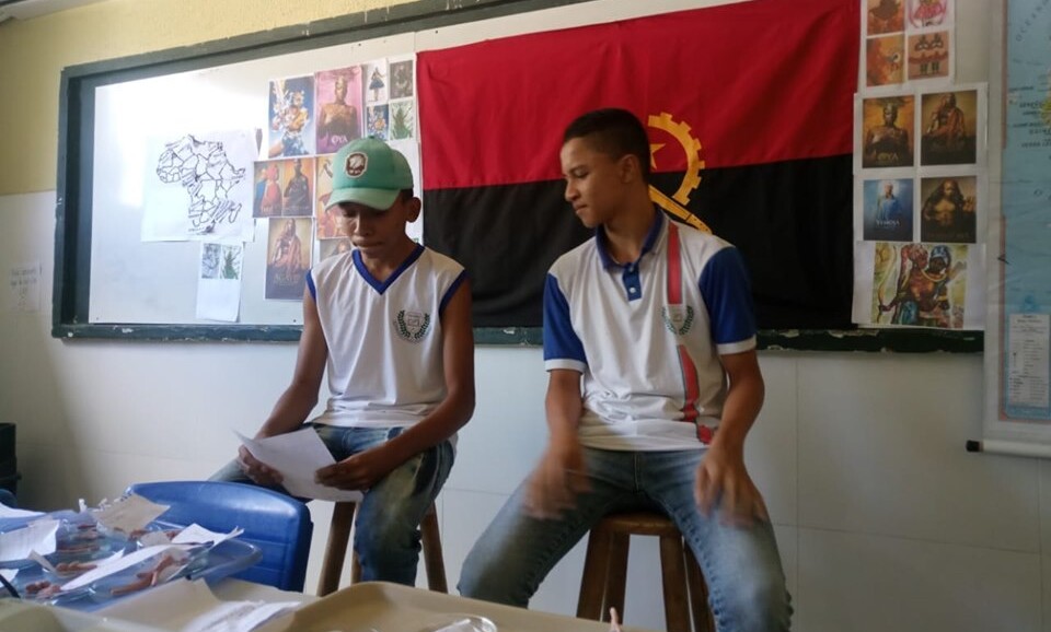 Foto em que há dois jovens sentados em banquetas. Eles estão na parte da frente de uma sala de aula apresentando um trabalho. Na lousa, atrás deles, há a bandeira da Angola - ela é dividida, horizontalmente numa metade superior vermelha e na outra parte inferior preta e no centro, há a roda dentada, a catana e a estrela em amarelo. Ao lado da bandeira, estão coladas imagens e um mapa do continente africano.