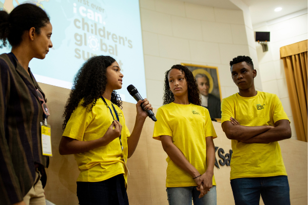Foto de uma apresentação, em que hpa três adolescentes de camiseta amarela à direita e uma adulta à esquerda. Uma das adolescentes está com microfone na mão falando.