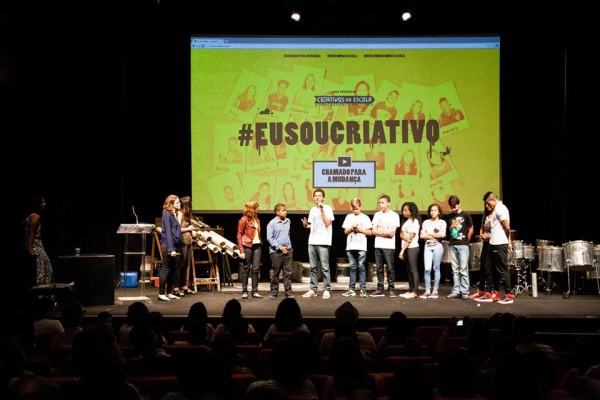 Foto de crianças em cima de um palco de teatro. Ao fundo, há um telão escrito 