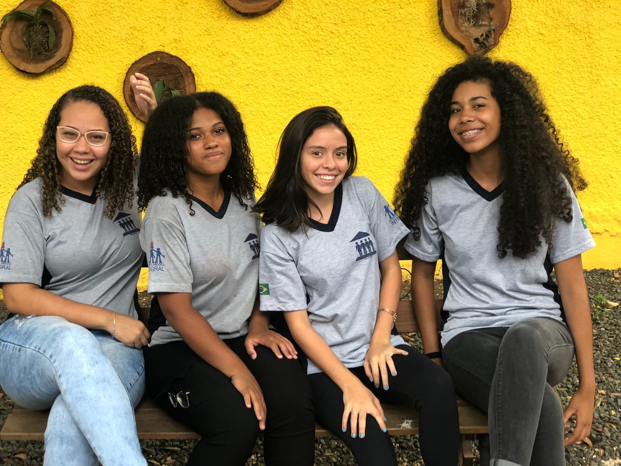 Foto de quatro adolescentes sentadas sorrindo para a foto. Elas usam uniforme escolar e ao fundo há uma parede amarela.