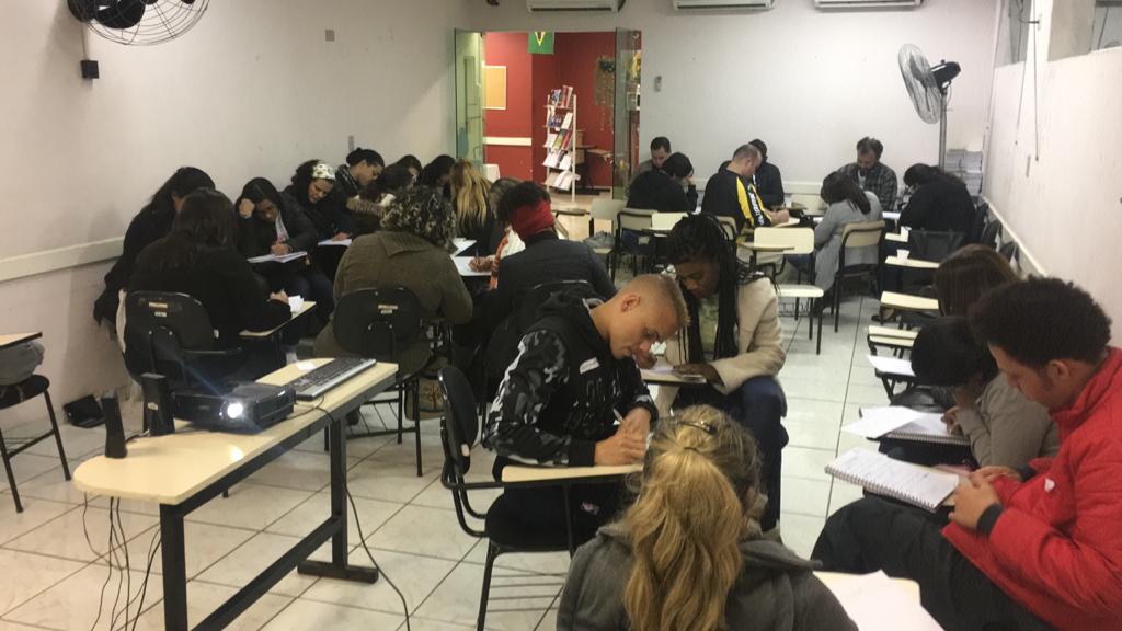 Foto de pessoas sentadas em carteiras numa sala de aula realizando atividade escrita.