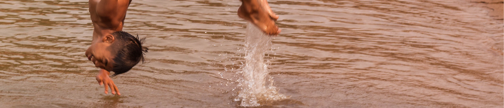 Foto de um adolescente indígena fazendo uma acrobacia ao pular em um rio. Ele sorri e está usando uma bermuda.