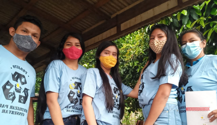 Foto de 5 pessoas em meio à floresta, utilizando camisetas azuis com um símbolo de reciclagem e máscaras de proteção. Há um homem, três adolescentes e uma mulher indígenas.