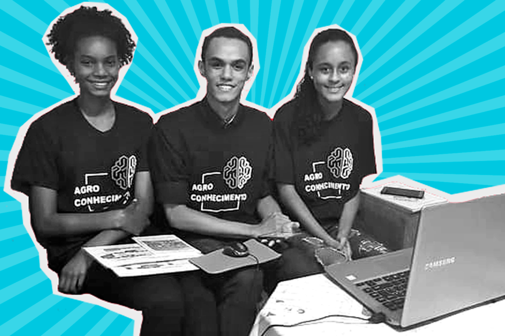 Foto em preto e branco de três adolescentes sentados em frente para um notebook - duas meninas de cabelos curtos e cacheados, e um menino - num fundo azul. Todos usam camiseta com o desenho de um cérebro e o nome “Agro Conhecimento”.