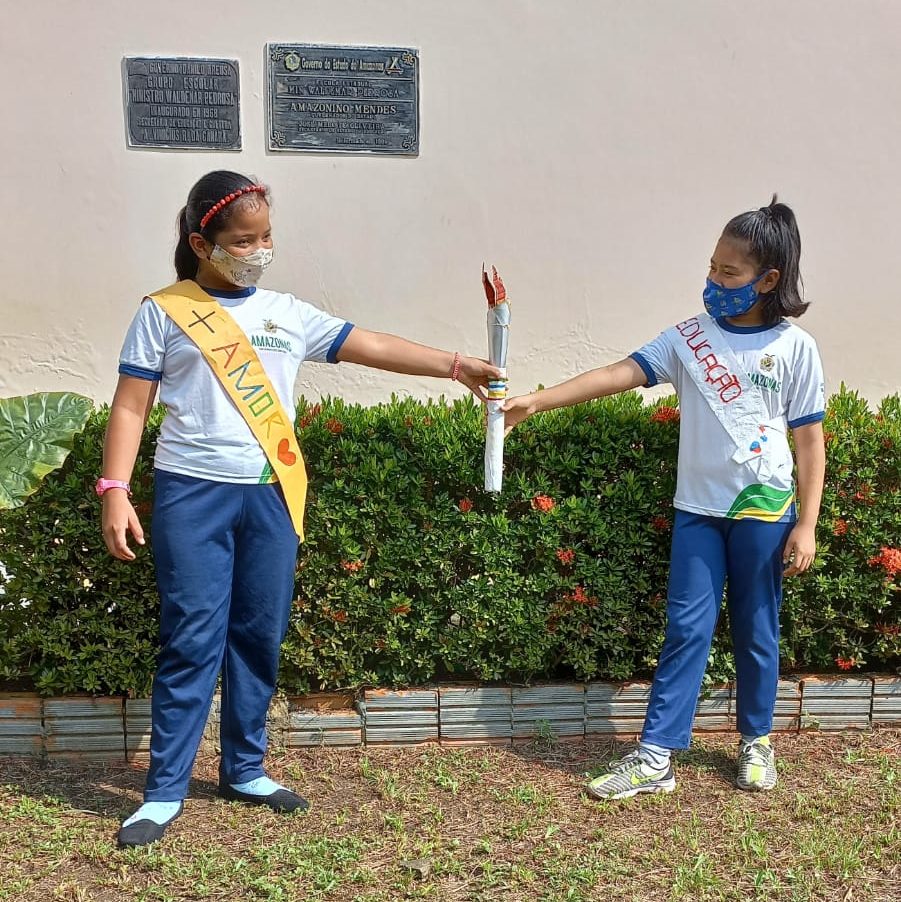 Duas meninas indígenas usam uniforme escolar e seguram uma tocha de papel. Elas usam ainda uma faixa cruzando o peito: uma com o dizer "+ amor" e outra com "educação. Elas estão em um jardim.