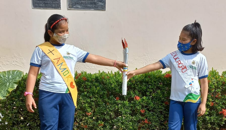 Duas meninas indígenas usam uniforme escolar e seguram uma tocha de papel. Elas usam ainda uma faixa cruzando o peito: uma com o dizer 