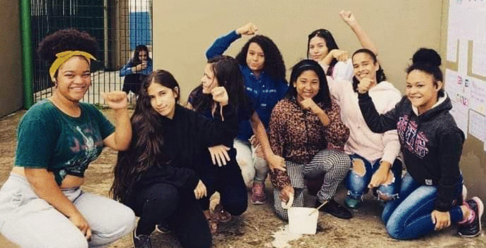 foto de um grupo de meninas - negras e brancas - agachadas e levantando os punhos fechados.