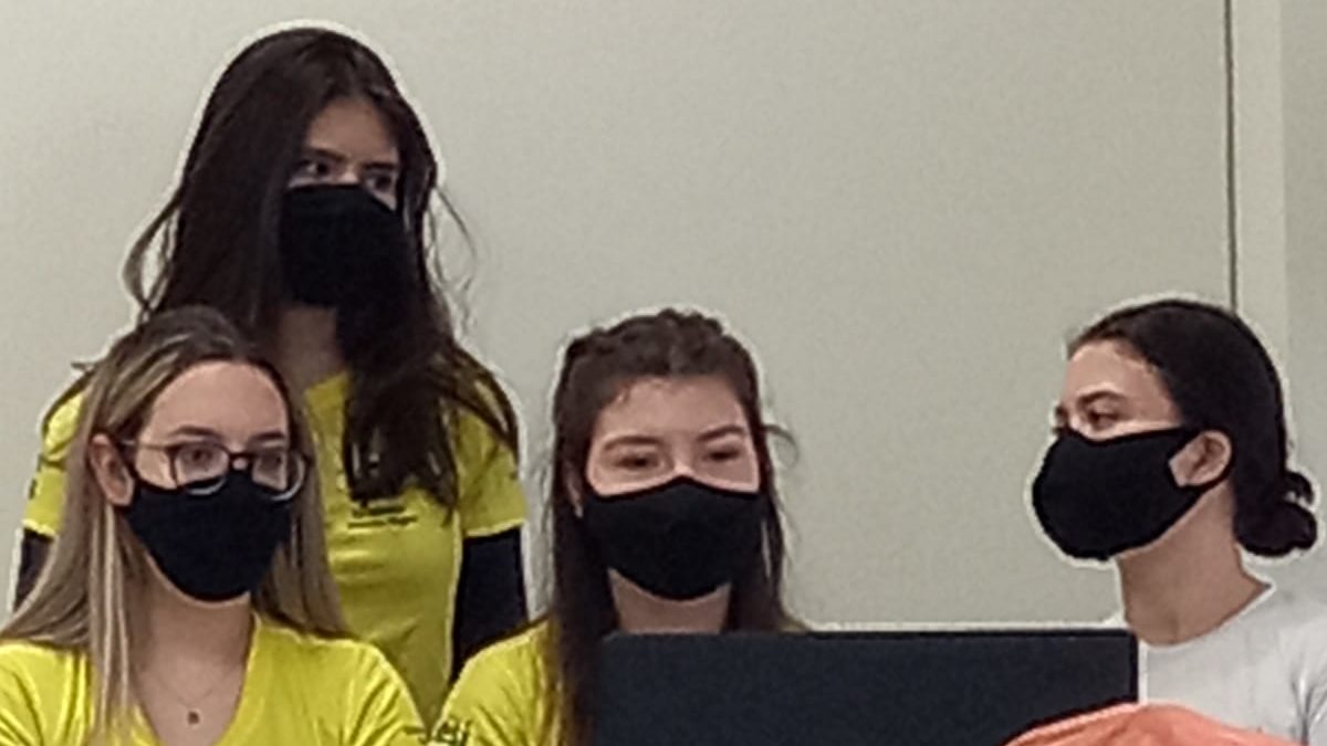 Quatro jovens brancas com máscaras de proteção pretas apresentam um trabalho em sala de aula