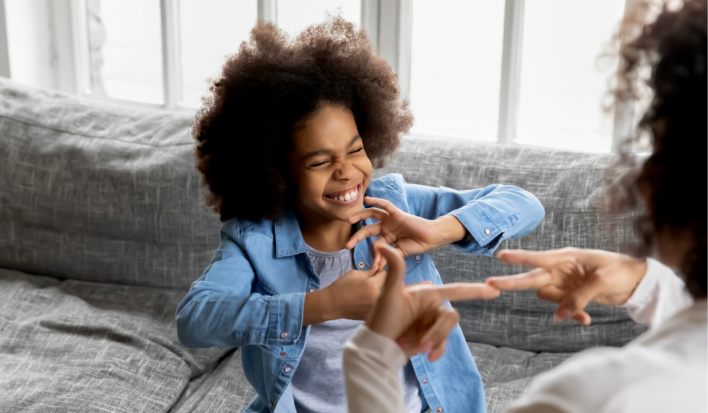 criança negra aprende língua de sinais em uma sala de estar, ela sorri.