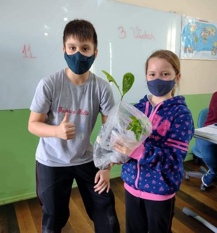 foto de duas crianças, uma segura uma planta e a outra faz um joia com a mão. Os dois usam máscara
